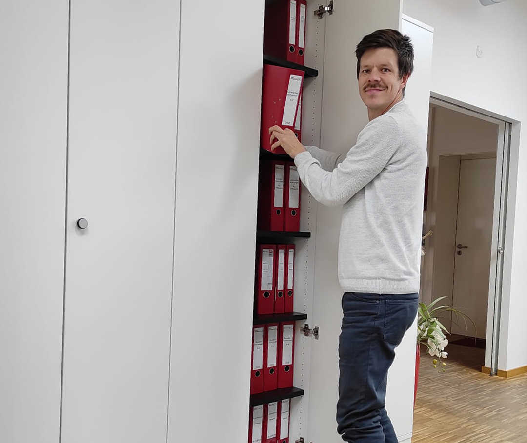 Das Bild zeigt Ricky stehend vor einem Aktenschrank Das Bild zeigt Ricky stehend vor einem Aktenschrank