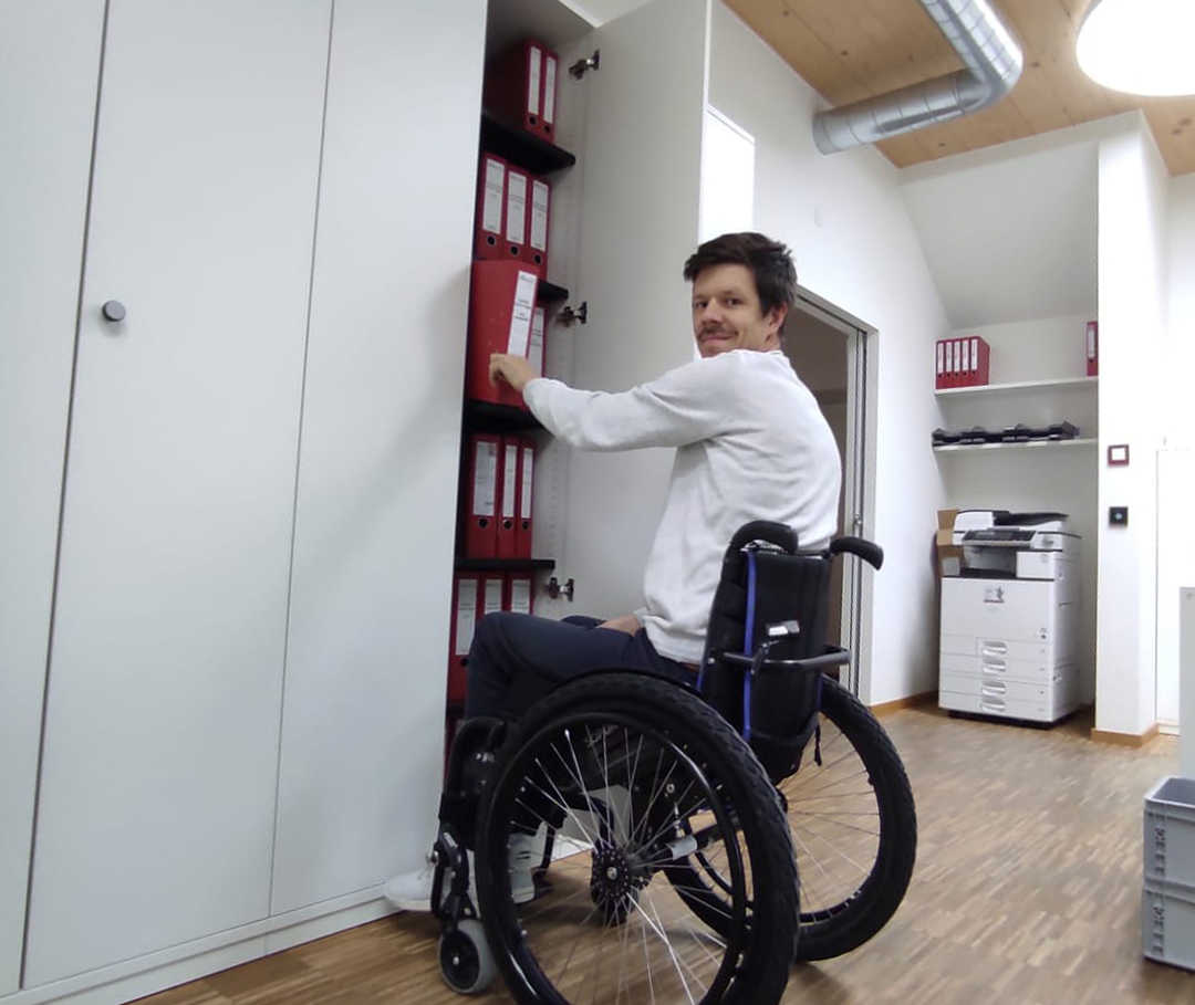 Das Bild zeigt Ricky im Rollstuhl vor einem Aktenschrank Das Bild zeigt Ricky im Rollstuhl vor einem Aktenschrank